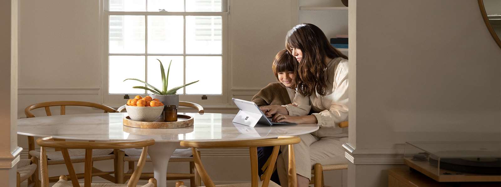 一位女性坐在餐桌旁，她的儿子在玩 Surface Go 2